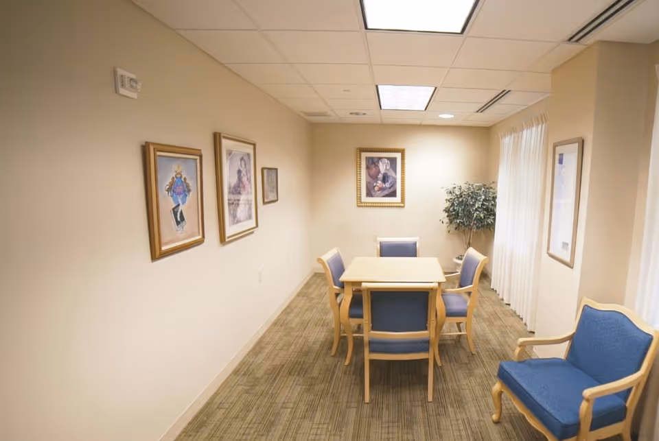 Small dining/meeting room with a square table surrounded by blue-upholstered chairs, framed artwork on the walls, and a potted plant.