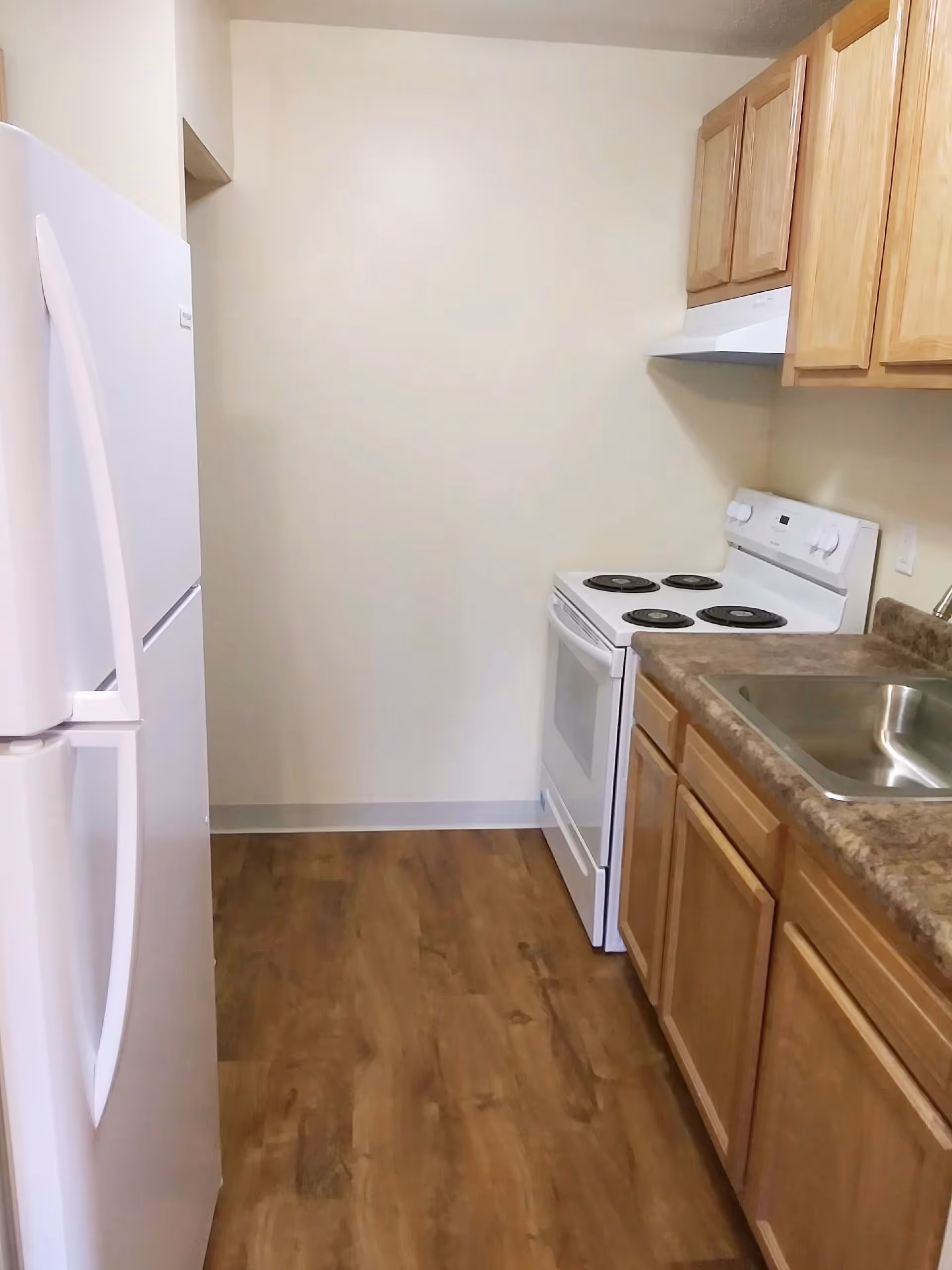 Narrow kitchen with a white refrigerator and electric stove, wooden cabinets, laminate countertops, and a stainless steel sink.