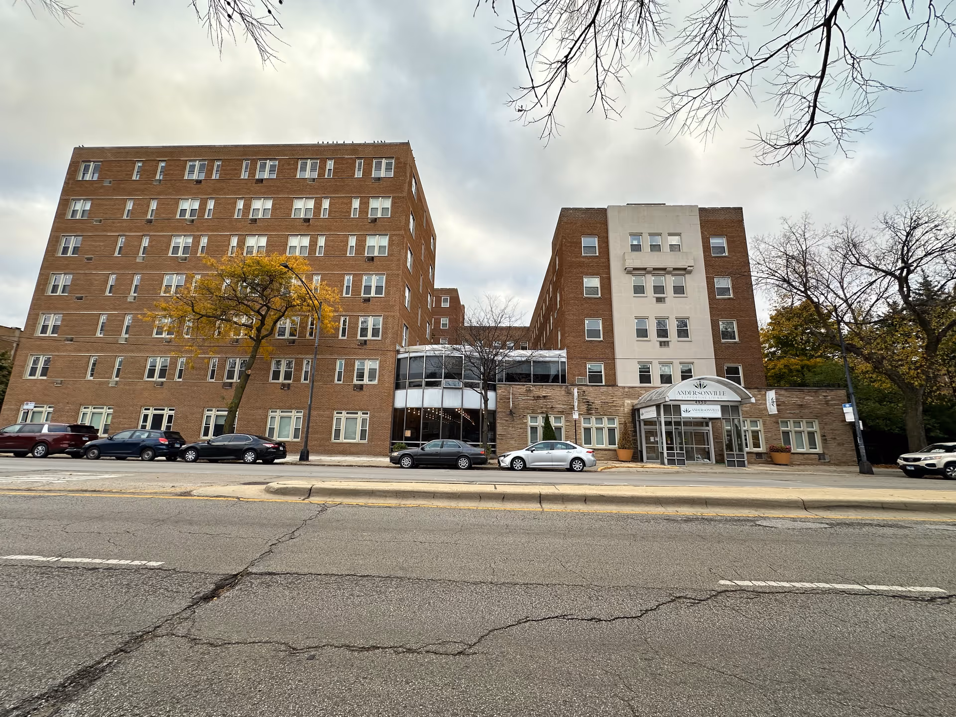 Exterior view of a large multi-story brick building with many windows, identified as Andersonville Senior Living. Several cars are parked along the street in front of the building, and leafless trees are visible around the area under a cloudy sky.