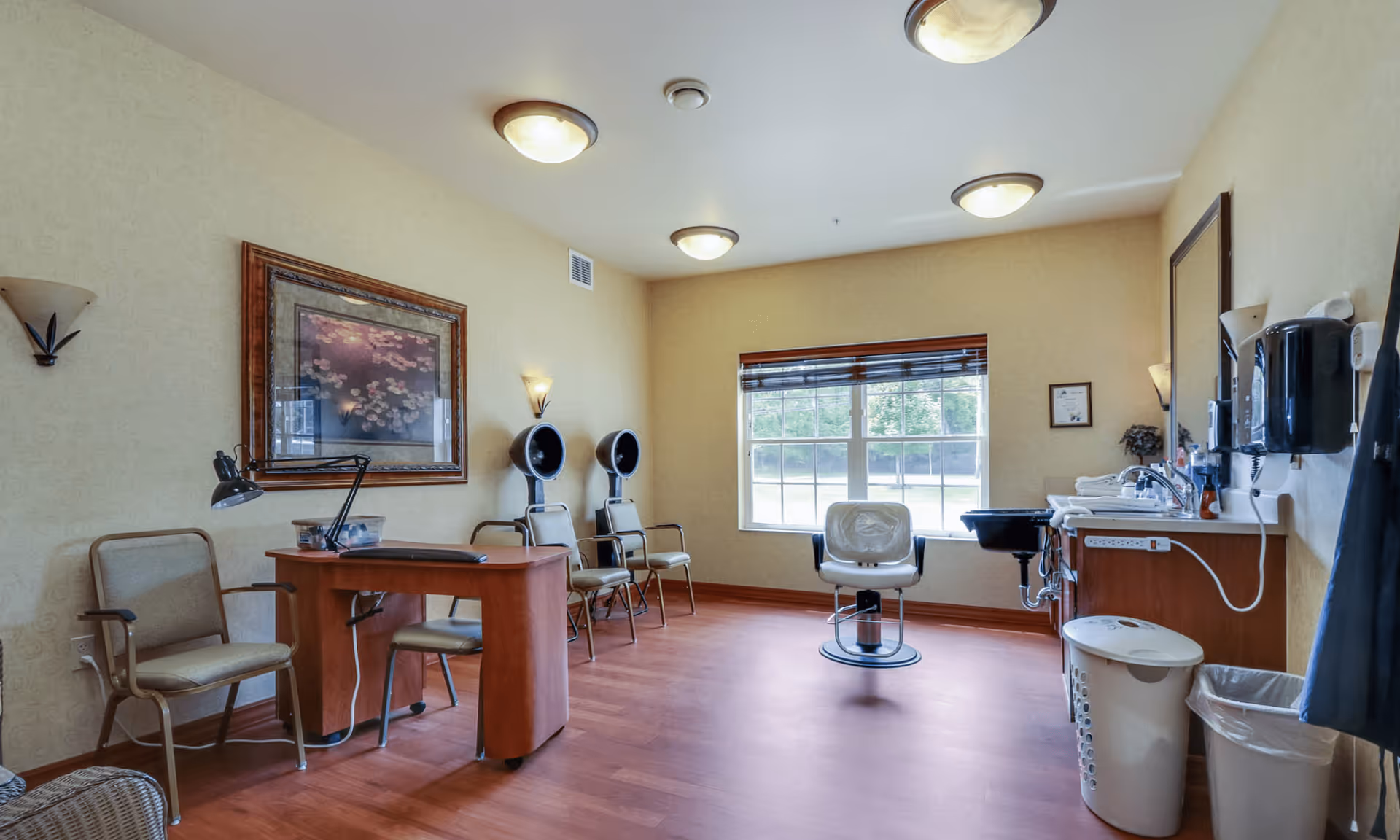 A well-lit room with wooden flooring and beige walls featuring a salon setup. There are two hair drying chairs with hooded dryers, a salon chair in front of a window with blinds, a small desk with a lamp and chairs, and a sink with hair washing facilities. The room has ceiling lights and wall sconces, and a framed picture on the wall.