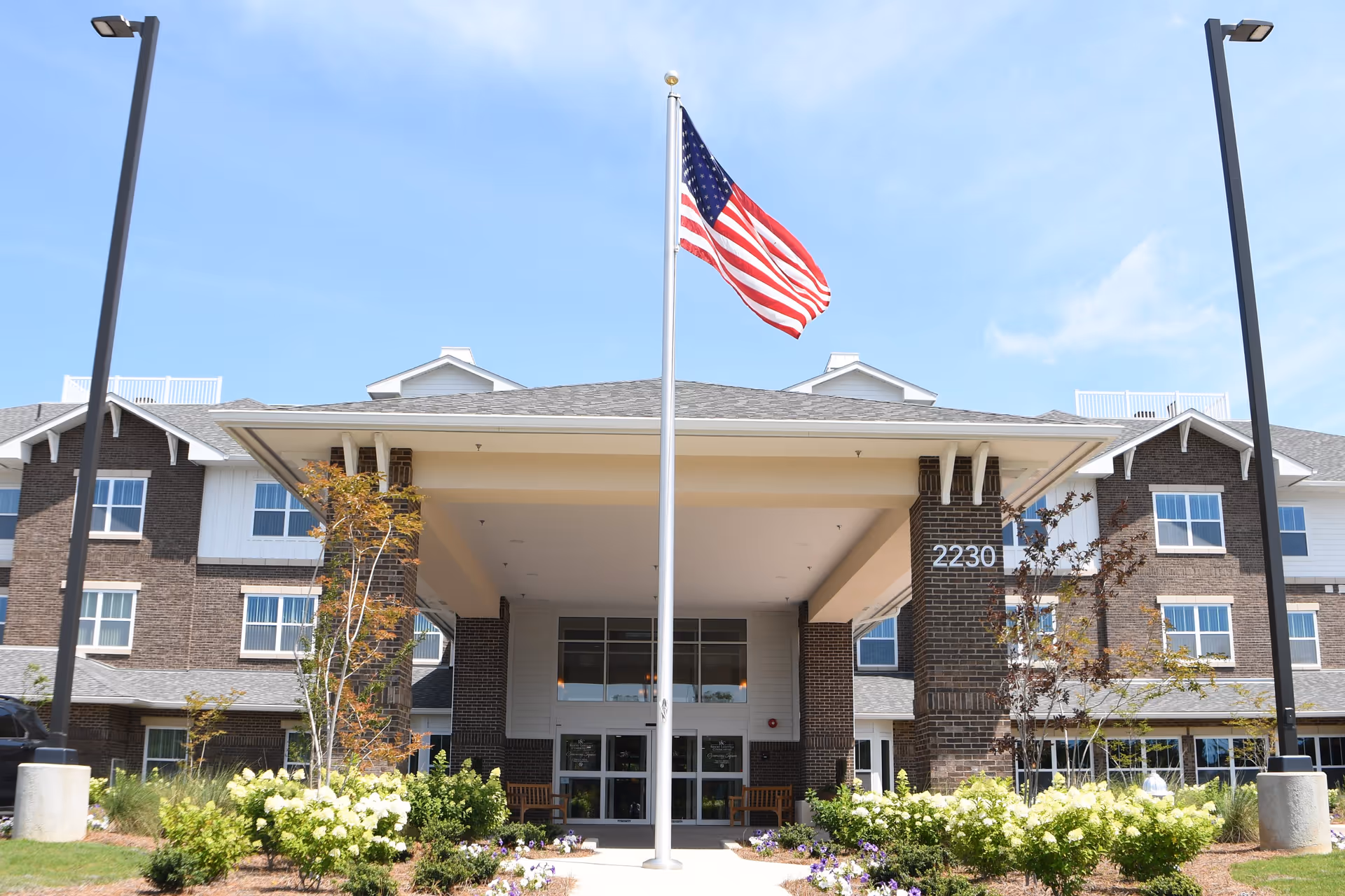 Front exterior view of Greenway Square Retirement Community building with an American flag on a flagpole in the center, surrounded by landscaped bushes and flowers, under a clear blue sky.