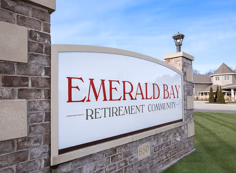 Brick monument entrance sign reading "Emerald Bay Retirement Community" beside a driveway with buildings in the background.