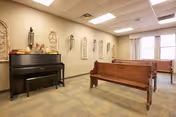 Interior room with wooden pew-style benches arranged facing a piano against the wall. The walls are decorated with ornamental wall hangings and the room is lit by ceiling lights. There are windows with curtains on one side.