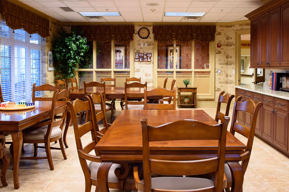 A warm and inviting dining room with several wooden tables and chairs arranged neatly. Large windows with blinds allow natural light to fill the room. The walls have a patterned wallpaper, and there is a clock and some framed pictures on the far wall. A small potted plant sits on a side table, and wooden cabinetry with a granite countertop is visible on the right side.