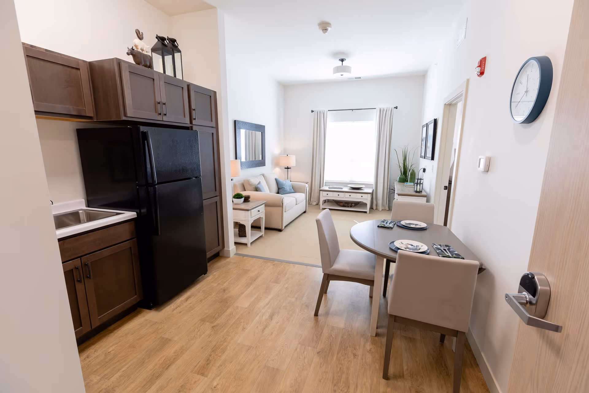 A bright and clean senior living apartment interior featuring a small kitchen area with dark wood cabinets and a black refrigerator on the left. Adjacent to the kitchen is a dining area with a round table set for two with plates and napkins. Beyond the dining area is a cozy living room with a beige sofa, a side table with a lamp, a large window with curtains, and a coffee table. The walls are white, and the floor is light wood. A clock is mounted on the wall near the entrance door on the right.