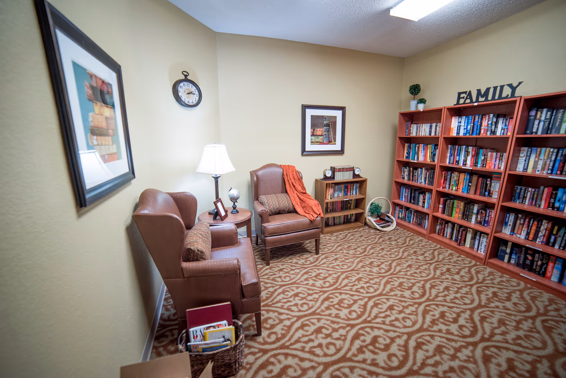 A cozy reading room with two brown armchairs, a small round wooden table with a lamp and a globe, a bookshelf filled with books, and a carpet with a decorative pattern. The wall has framed artwork and a clock, and there is a basket with magazines on the floor. The word FAMILY is displayed on top of the bookshelf.