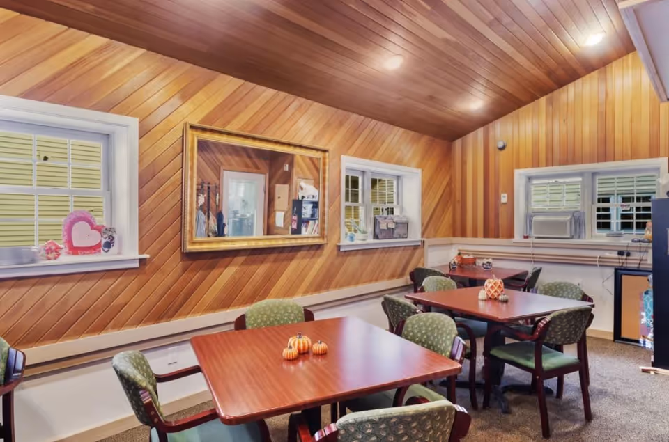 Interior room with wooden paneled walls and ceiling, featuring three tables each surrounded by green cushioned chairs. Small decorative pumpkins are placed on the tables. There are three windows with white frames, one with an air conditioning unit, and a large gold-framed mirror on the wall.