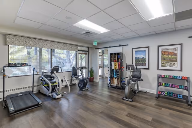 A fitness room with exercise equipment including a treadmill, stationary bikes, and a rack of colorful dumbbells. The room has large windows with blinds, framed artwork on the walls, and wood-style flooring.