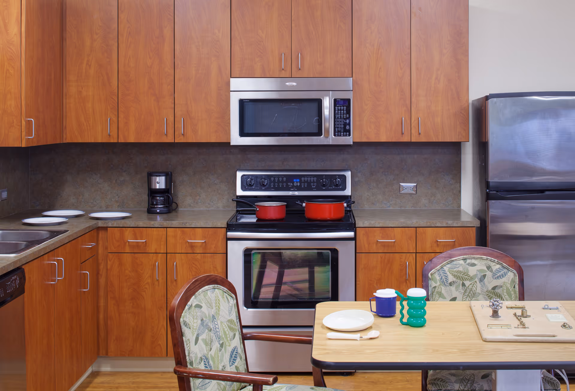 A kitchen area with wooden cabinets, a stainless steel microwave and oven, a coffee maker, and a stainless steel refrigerator. There is a table with two chairs featuring leaf-patterned upholstery. On the table are a plate, a spoon, a cup, a green water bottle, and a small activity board.