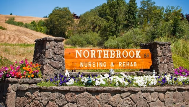 A wooden sign mounted on a stone structure surrounded by colorful flowers. The sign reads 'NORTHBROOK NURSING & REHAB' with the number 64 on the right side. The background shows grassy hills and trees under a clear blue sky.