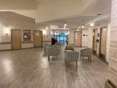 A spacious interior common area with light wood flooring and walls. There are six striped armchairs arranged in a circle in the center of the room. The area is well-lit with ceiling lights and wall sconces. Several doors and a bulletin board are visible along the walls, and a glass exit door is seen at the far end.