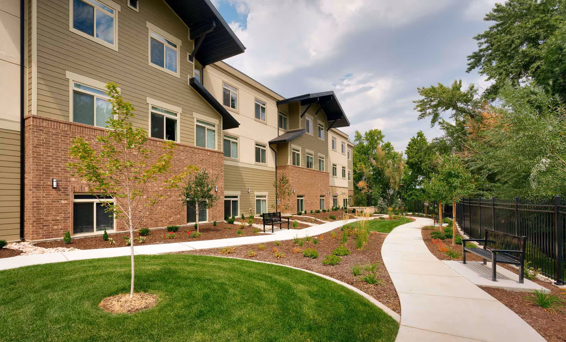 Three-story senior living building with a landscaped courtyard, curved walkway, benches, and young trees.