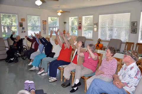 A group of elderly people sitting in chairs arranged in a semi-circle in a bright room with large windows. Most of them have their arms raised, participating in a group activity. The room has light-colored walls and ceiling fans, with some framed pictures and plants visible.