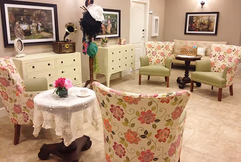 A cozy sitting area with floral upholstered armchairs arranged around small wooden tables. One table is covered with a white lace tablecloth and has a small flower arrangement. The room has light-colored walls adorned with framed paintings, a coat rack with hats, and cream-colored dressers along the wall.