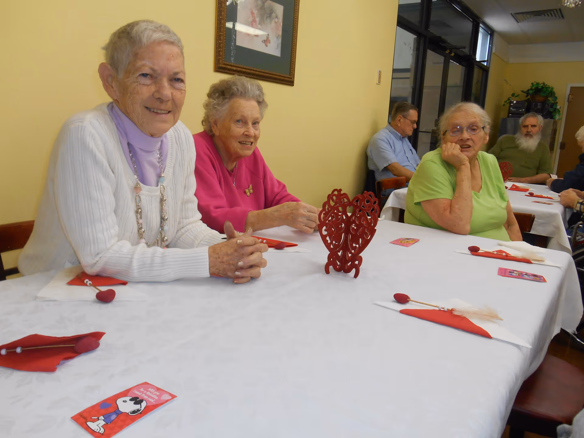 A group of elderly people sitting around a table in a senior living facility dining room. The table is covered with a white tablecloth and decorated with red napkins and a red heart-shaped centerpiece. The walls are painted yellow, and there is a framed picture hanging on the wall.