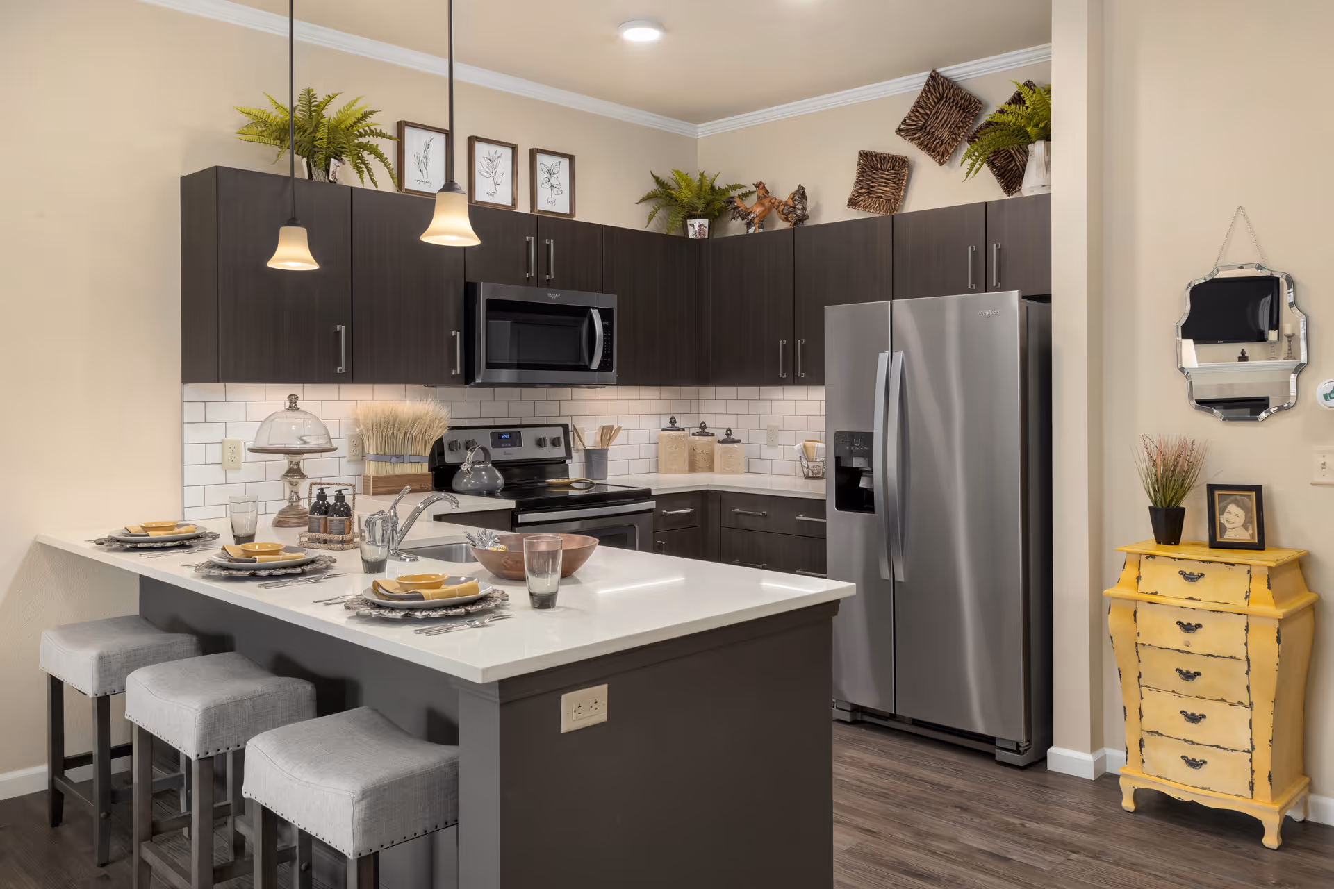 Modern kitchen with dark wood cabinets, white countertops, and stainless steel appliances including a refrigerator and microwave. There is a kitchen island with three gray cushioned stools and place settings for three. The backsplash is white subway tile, and there are decorative plants and framed artwork on top of the cabinets. A small yellow chest with a plant and framed photo sits against the wall next to a decorative mirror.