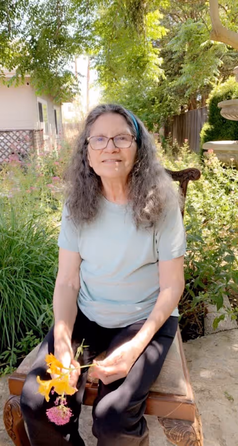 An elderly woman with long gray hair and glasses is sitting on a wooden bench outdoors in a garden. She is wearing a light blue t-shirt and black pants, holding a small bouquet of flowers. The background shows green plants, trees, and part of a house.
