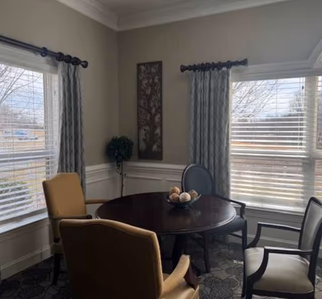 A cozy interior room with a round dark wooden table surrounded by four chairs, two with beige upholstery and two with light gray upholstery. The room has two large windows with white blinds and patterned gray curtains. A decorative wall art piece and a small potted plant are placed in the corner.