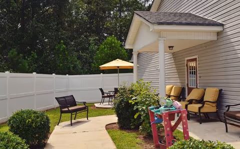 Outdoor patio area at a senior living facility with cushioned chairs, a small table with an umbrella, a white privacy fence, and green bushes along a curved concrete pathway.
