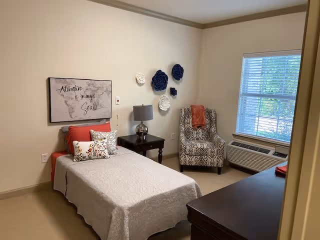 A neatly arranged bedroom with a single bed covered in a light gray bedspread and decorated with multiple pillows. Above the bed is a wall art piece with the text 'Adventure is my Soul'. Next to the bed is a dark wooden nightstand with a silver lamp. On the wall above the nightstand are decorative blue and white flower wall hangings. A patterned armchair with an orange throw blanket is placed near a window with blinds. Below the window is an air conditioning unit. A dark wooden dresser is partially visible in the foreground.