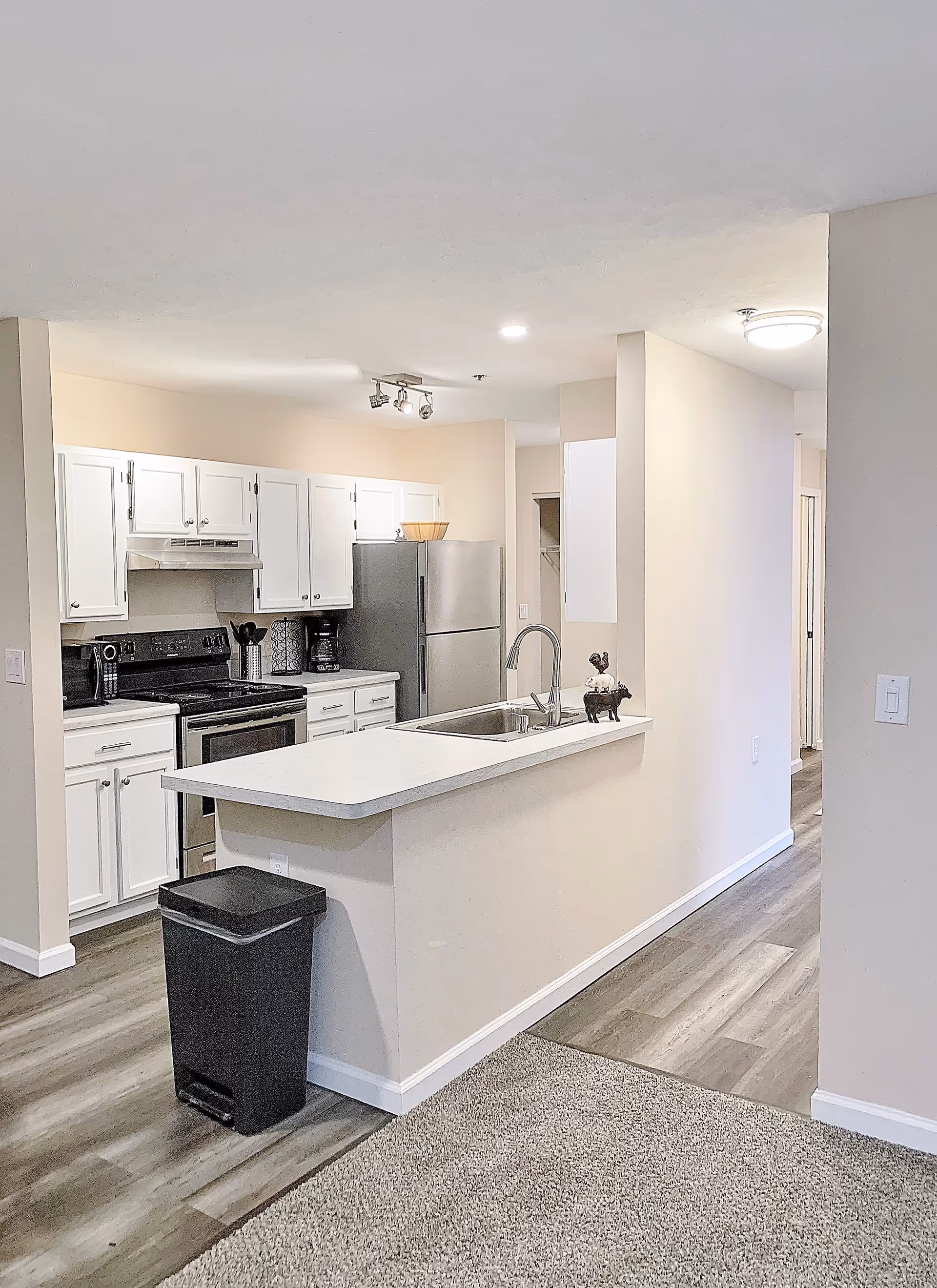 A modern kitchen area in a senior living facility featuring white cabinets, a stainless steel refrigerator, a stove with an oven, a coffee maker, and a countertop with a sink. The floor is a combination of wood and carpet, and there is a black trash bin next to the counter. The walls are painted in a light beige color, and the area is well-lit with ceiling lights.
