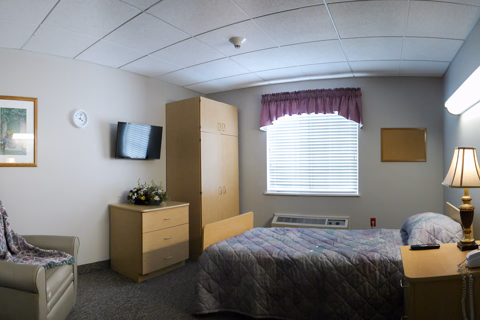 Single-occupancy bedroom with a bed, dresser, wall-mounted TV, armchair and a window with blinds.