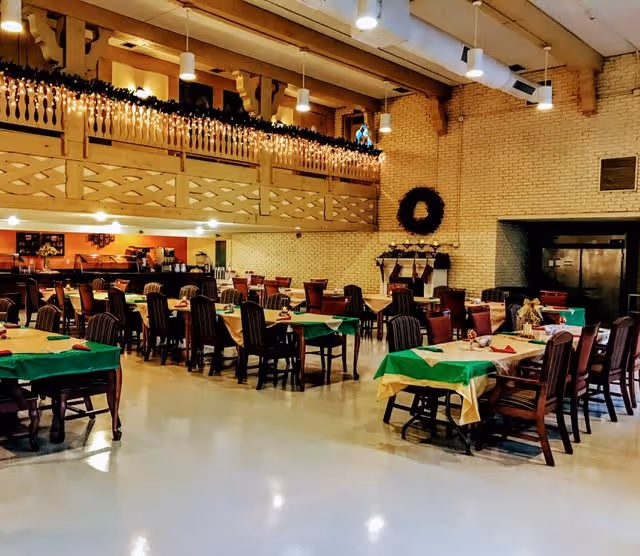 Spacious dining room with multiple tables covered in green and yellow tablecloths, surrounded by wooden chairs. The room features a high ceiling with exposed wooden beams, hanging lights, and a balcony railing decorated with garland. A large wreath and stockings are hung on a brick wall, suggesting a festive holiday setting.