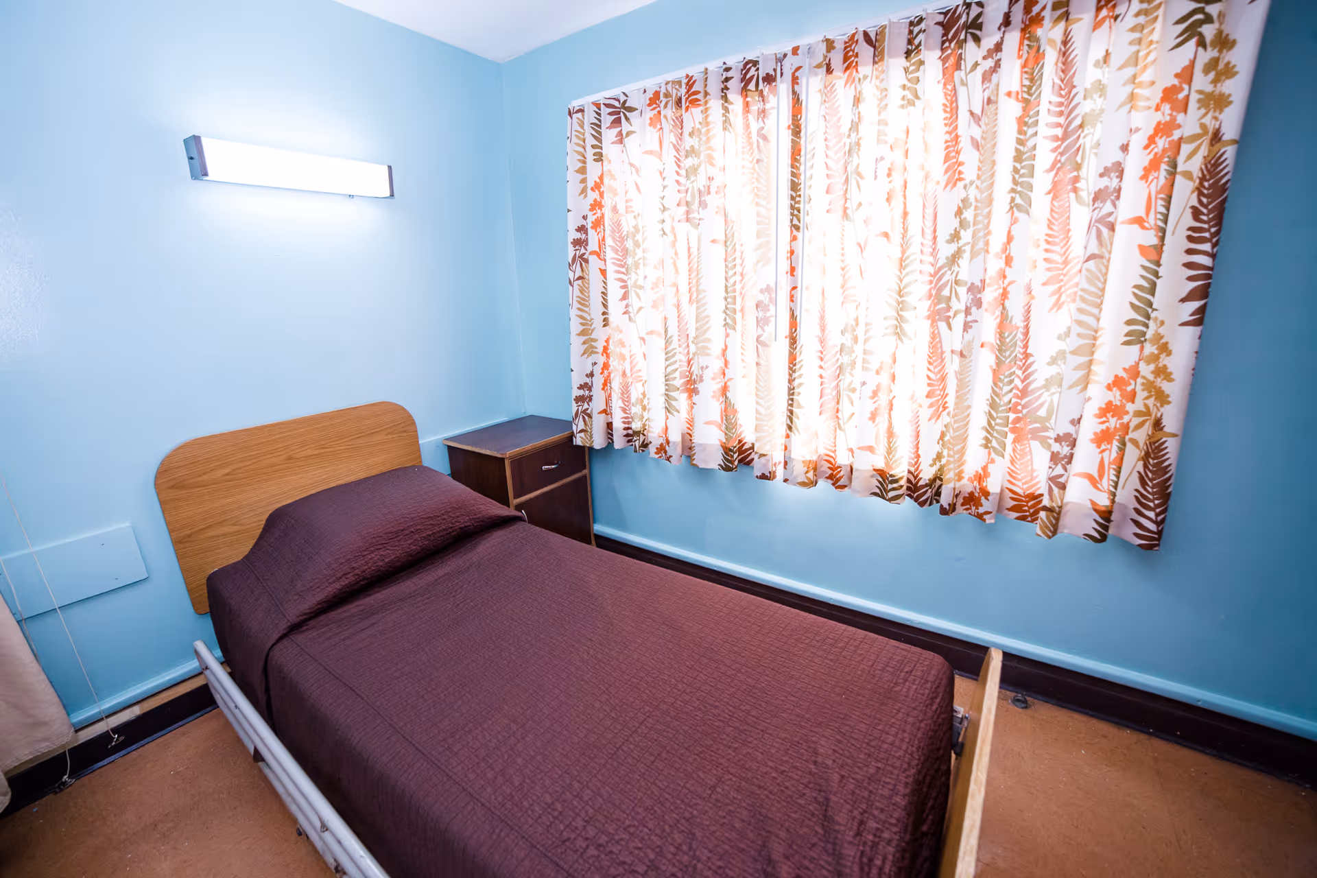 A small bedroom with light blue walls, a single bed with a brown bedspread, a wooden headboard, a small wooden nightstand with two drawers, and a window with floral patterned curtains letting in bright light.