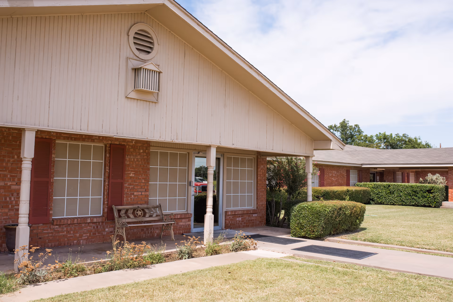Exterior view of a single-story brick building with white framed windows and red shutters. There is a covered porch area with a decorative bench and neatly trimmed bushes along the walkway. The sky is partly cloudy.