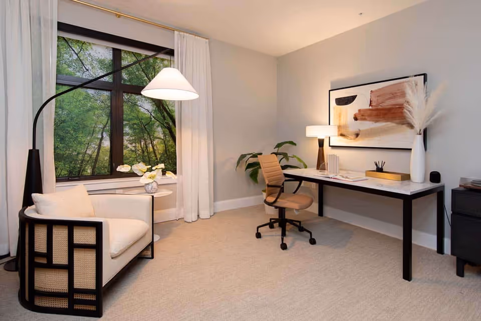 Bright furnished sitting area with an upholstered armchair by a large window and a desk with an office chair, lamp, and artwork on the wall.