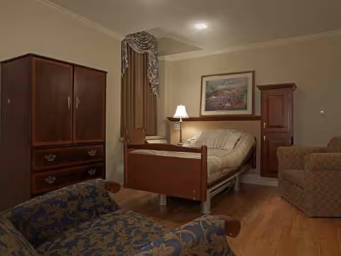 A cozy bedroom in a senior living facility with a wooden bed, armoire, lamp, and seating.