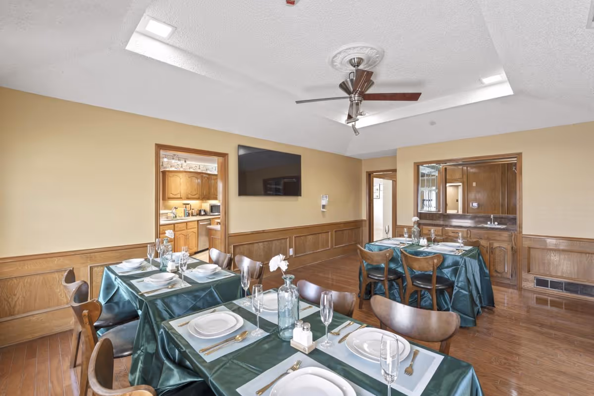 A dining room with three tables covered in green tablecloths, each set with white plates, glasses, and silverware. The room has wooden wainscoting, a ceiling fan, and a wall-mounted TV. There is a doorway leading to a kitchen with wooden cabinets and another doorway to an adjacent room.