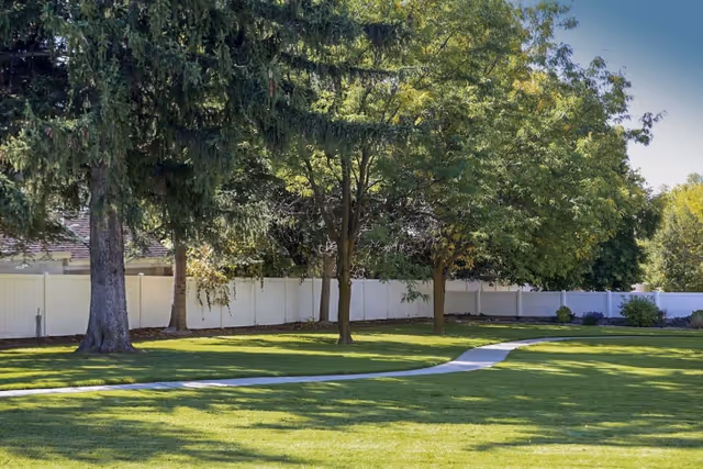 Grassy courtyard with a curved concrete path, mature trees, and a white privacy fence under a clear sky.