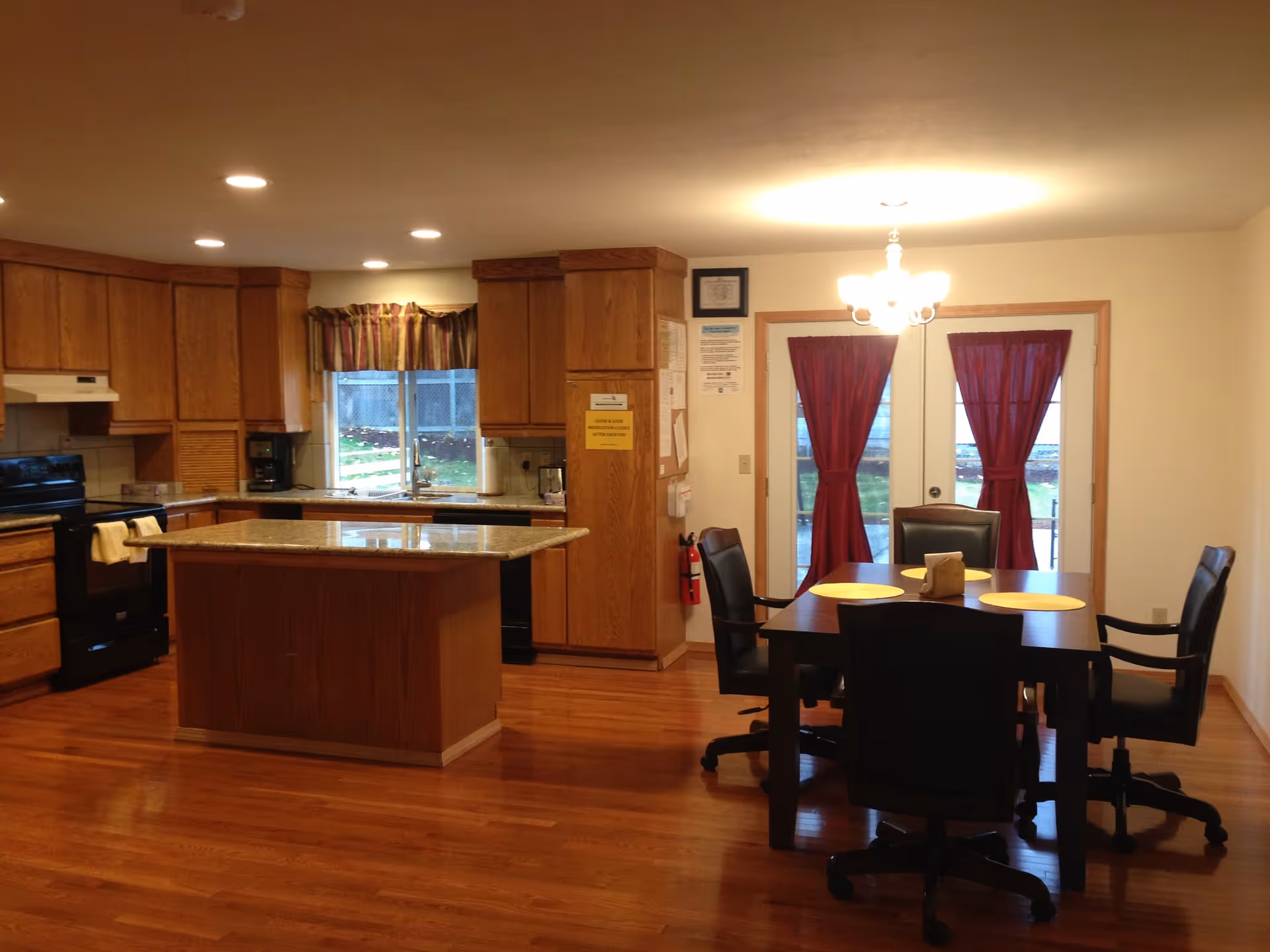 A spacious kitchen and dining area with wooden floors and cabinetry. The kitchen features an island with a granite countertop, black appliances including a stove and dishwasher, and a window with a valance above the sink. The dining area has a dark wooden table with four black chairs on wheels, yellow placemats, and a chandelier overhead. Red curtains cover the glass doors leading outside.