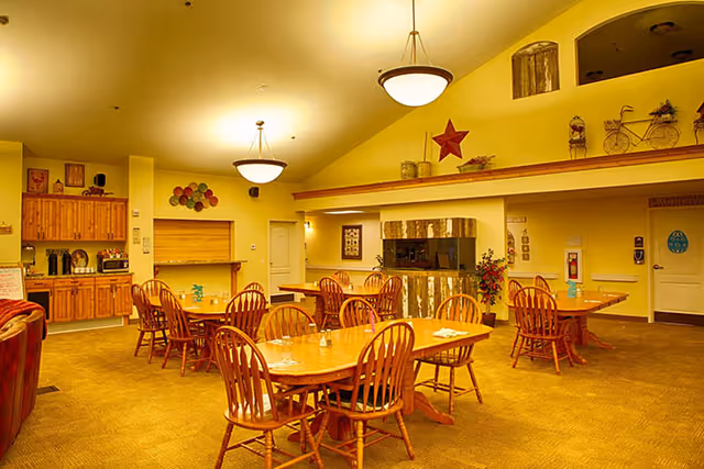 A warm, inviting dining room with wooden tables and chairs arranged neatly. The room features a rustic fireplace with a wooden mantel, decorative items on shelves above, and soft yellow lighting from ceiling fixtures. There is a kitchenette area with wooden cabinets and a coffee station on one side, and a door labeled 'Laundry' on the far wall.