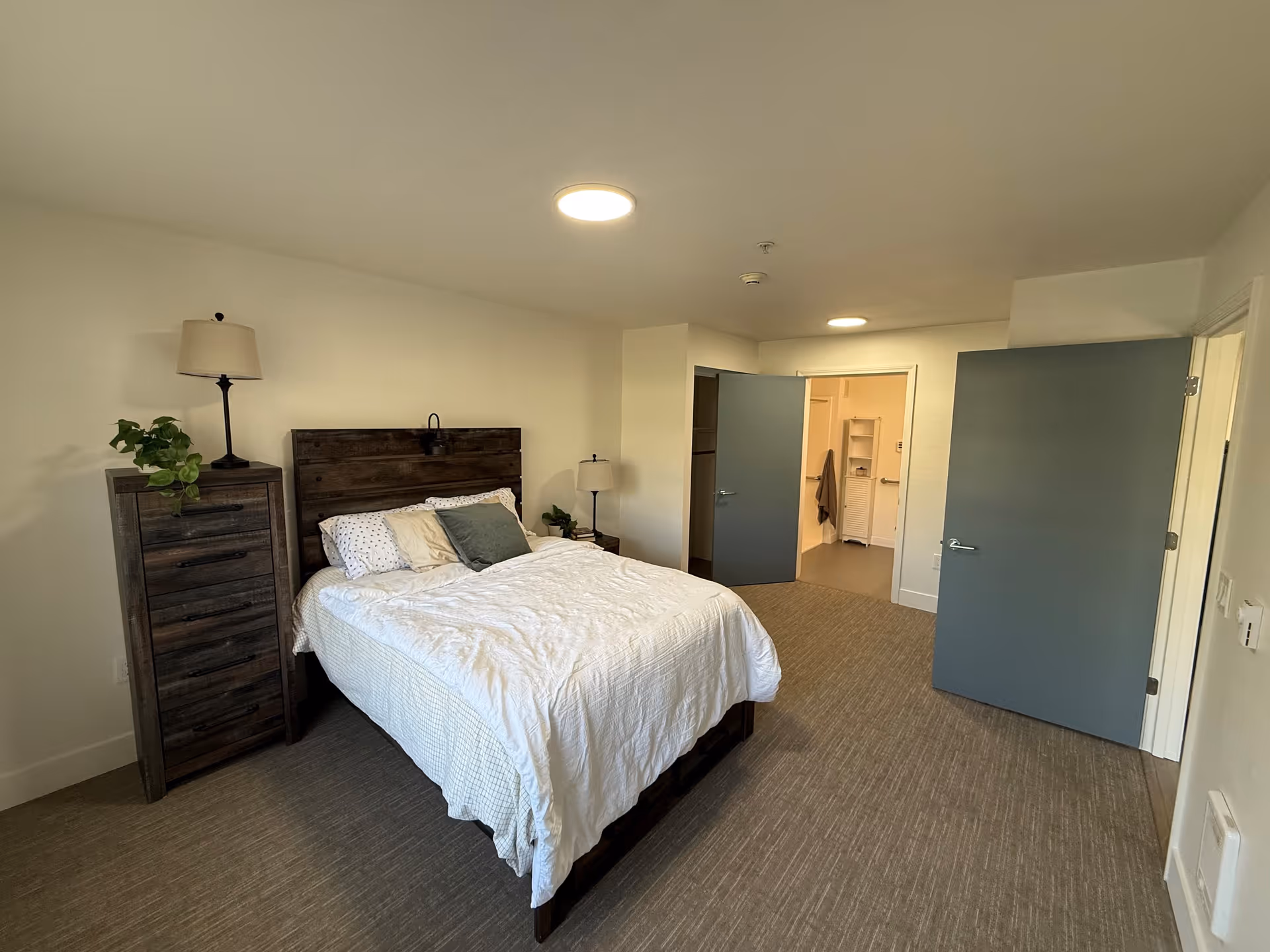 A bedroom with a neatly made bed featuring white bedding and multiple pillows. To the left of the bed is a tall wooden dresser with a lamp and a small plant on top. The room has beige carpet flooring and light-colored walls. Two blue doors are open, one leading to a bathroom area with visible shelving and a towel hanging, and the other leading to another part of the facility. The ceiling has two round recessed lights.