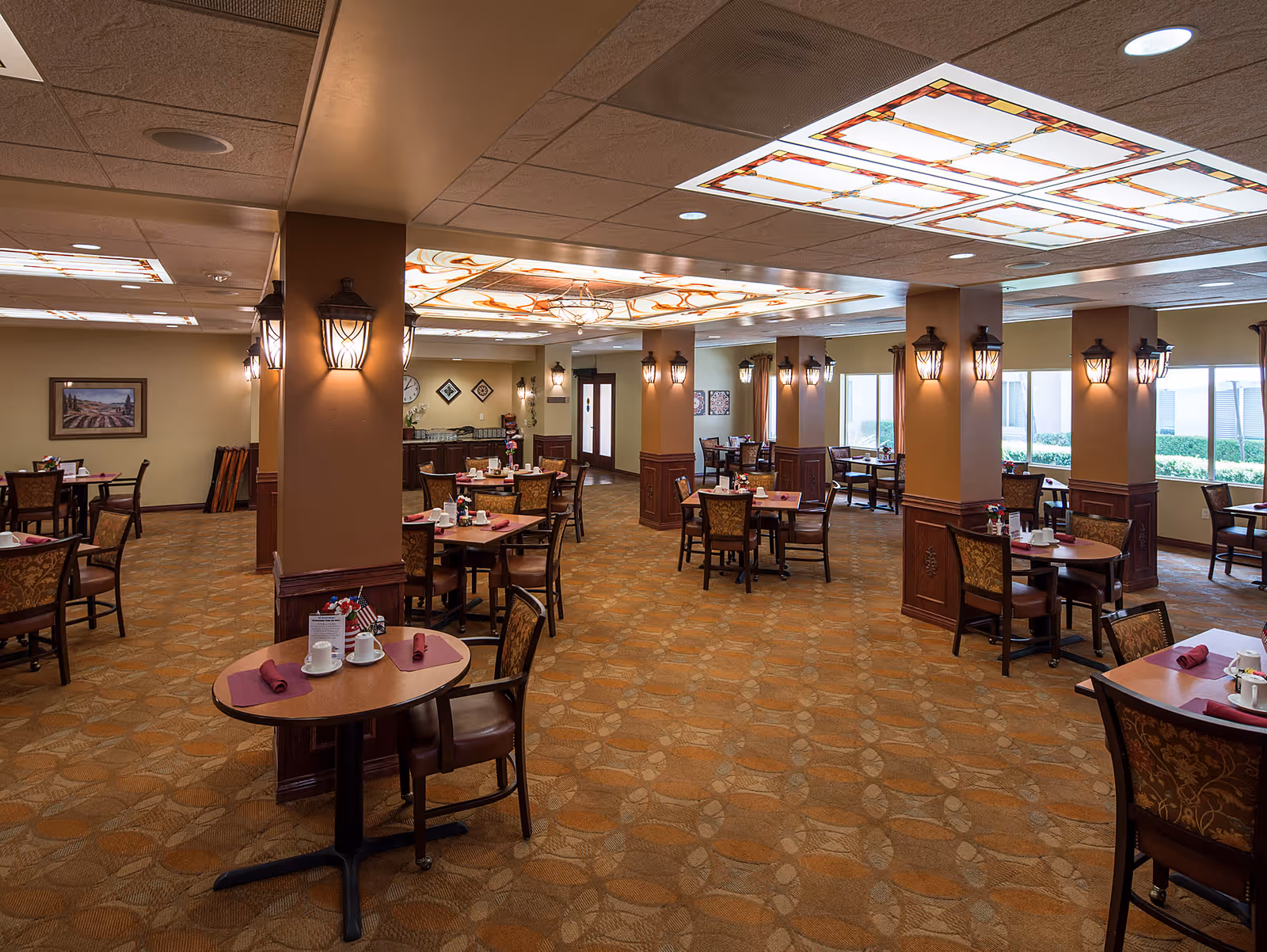 Spacious dining room with multiple round tables and chairs arranged neatly. The room features warm lighting from wall-mounted lantern-style fixtures and decorative ceiling lights. The carpet has a patterned design, and the walls are adorned with framed artwork. Large windows on one side allow natural light to enter the room.