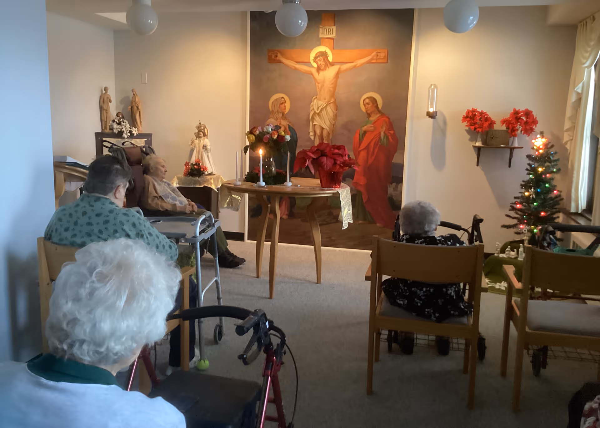 A small group of elderly people seated in a room facing a religious altar with a large painting of Jesus on the cross and two other figures. The room is decorated with candles, flowers, religious statues, and a small Christmas tree with lights. Some of the elderly individuals use walkers and wheelchairs.