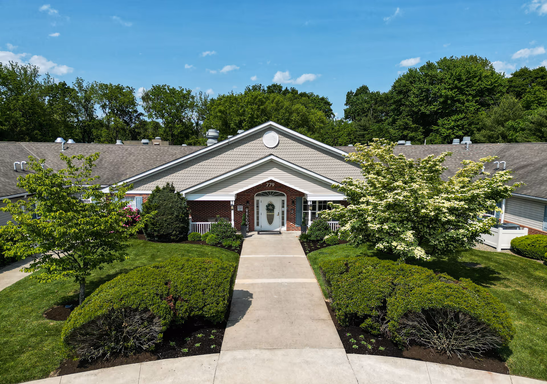 Front entrance of a single-story brick and siding senior care building with a central walkway, porch, and landscaped shrubs.