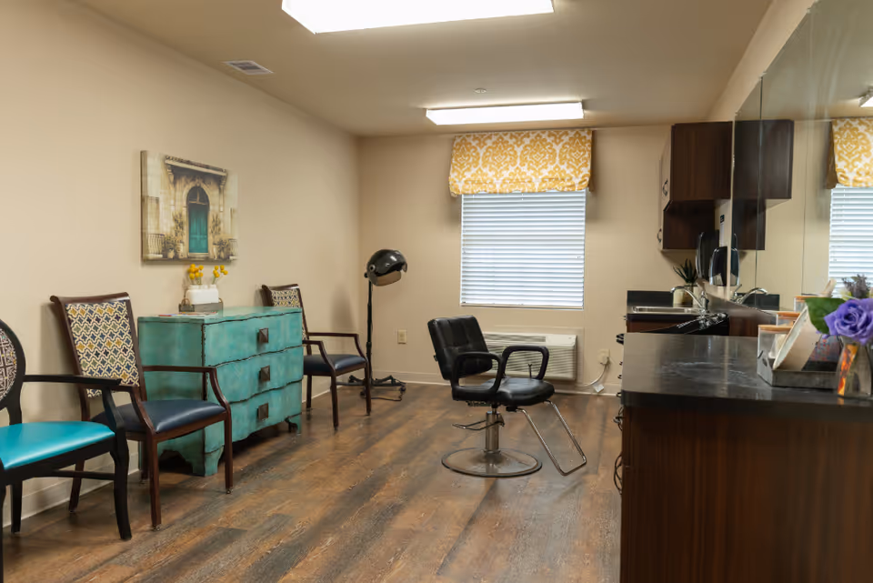 Interior room with a black salon chair in the center, a turquoise chest of drawers against the wall with three chairs around it, a hair dryer stand, and a countertop with a mirror and cabinets above it. The room has wood flooring, beige walls, a window with a yellow patterned valance, and a framed picture on the wall.