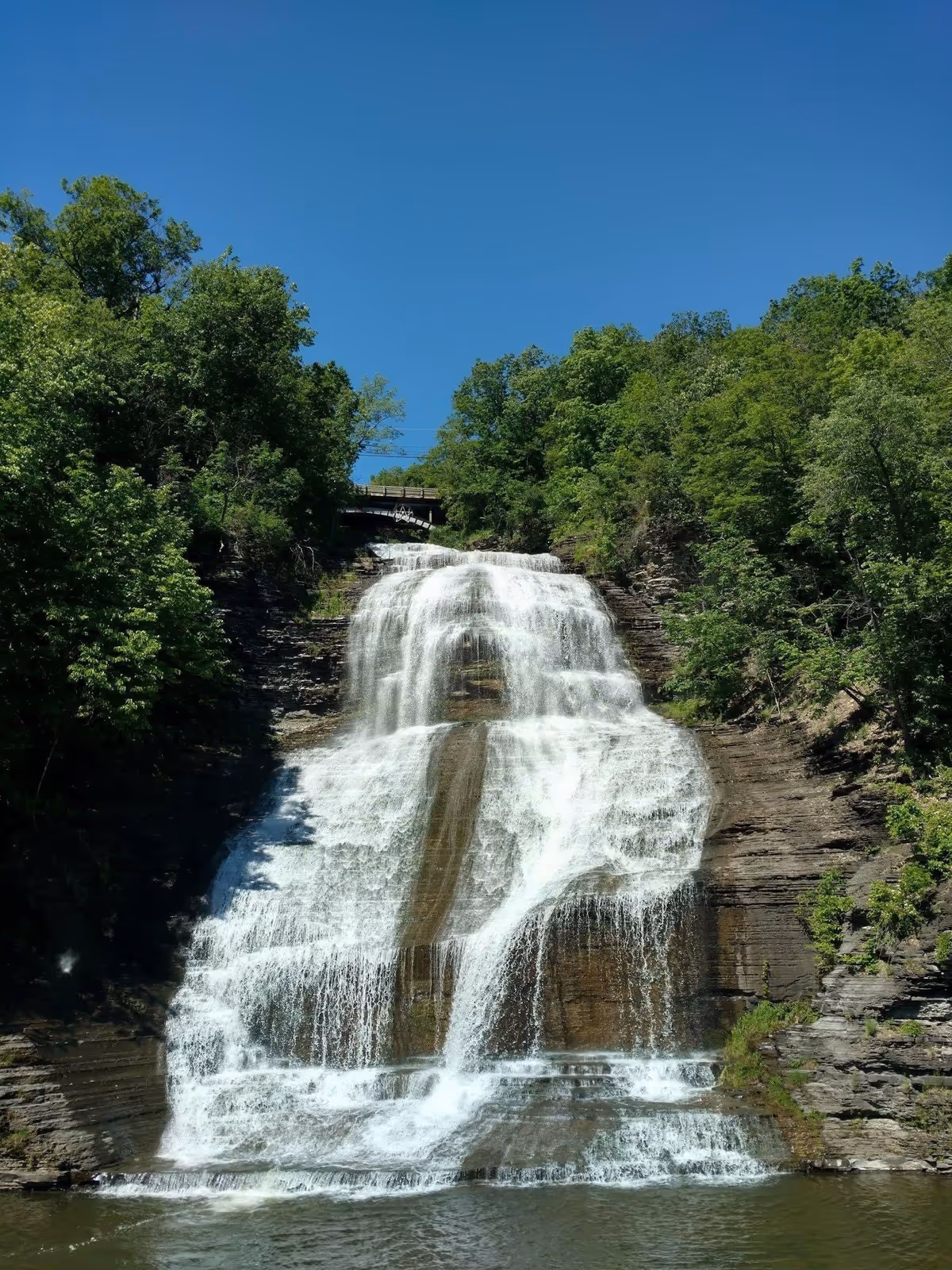 A large waterfall cascading down layered rock formations surrounded by lush green trees under a clear blue sky.