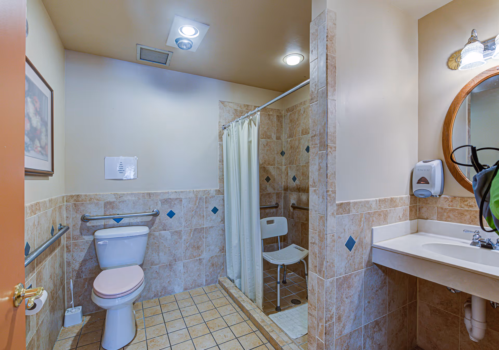 A bathroom with beige tiled walls and floor, featuring a toilet with grab bars on both sides, a walk-in shower with a white shower curtain and a white shower chair, and a sink with a round mirror above it. There is a soap dispenser mounted on the wall next to the sink and a framed picture on the wall near the door.