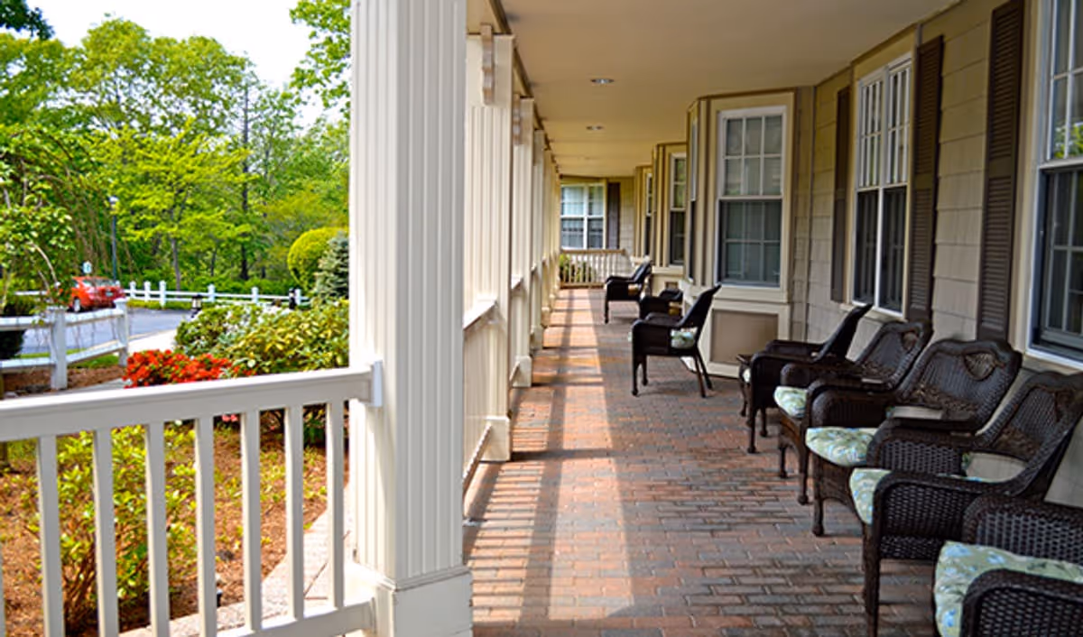 A covered outdoor porch area with several wicker chairs with cushions lined up along the wall of a building. The porch has brick flooring and white columns, overlooking a landscaped garden with green trees and bushes.