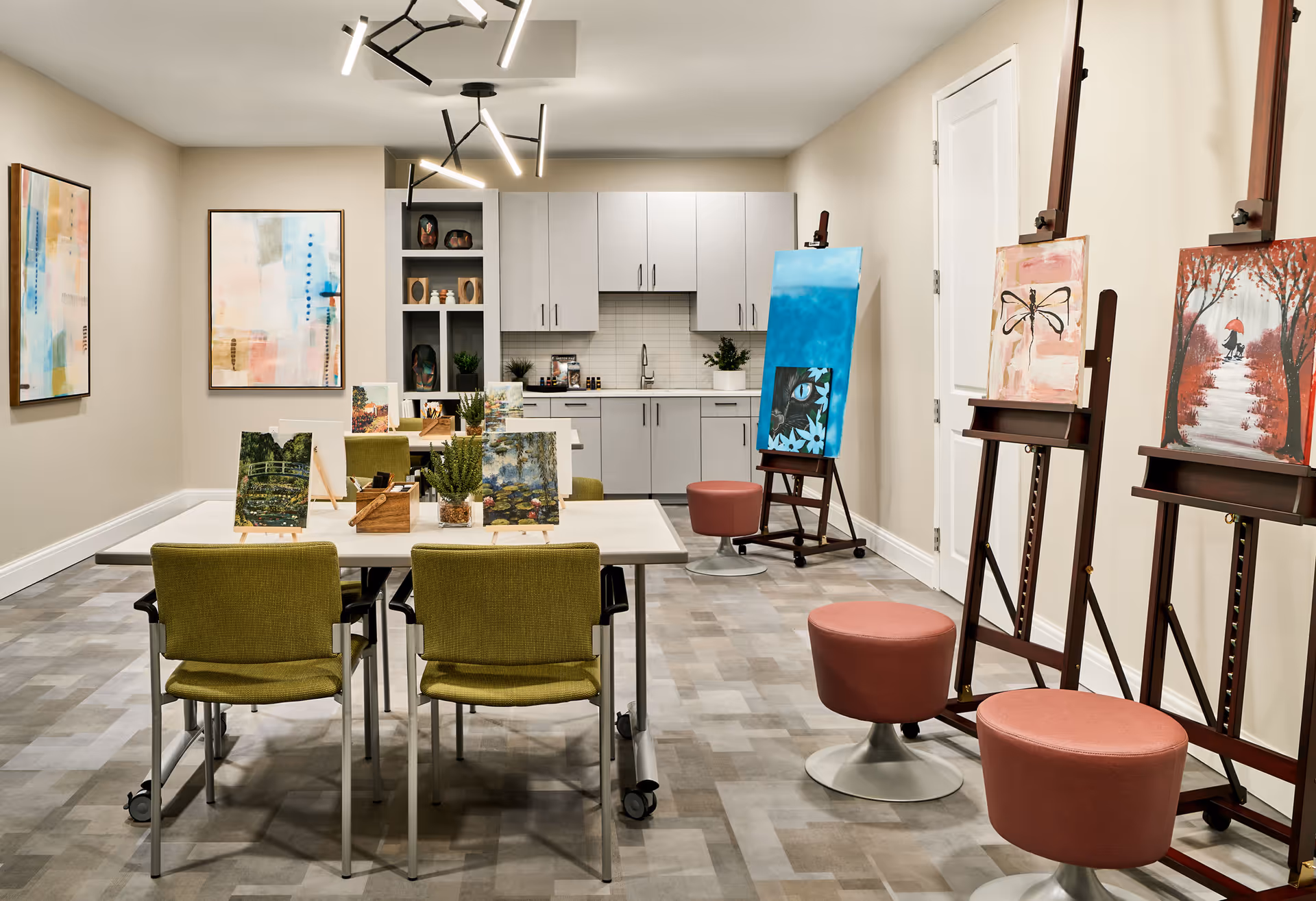 A bright art studio room with several paintings on easels and tables. The room features modern lighting, green chairs, and a kitchenette with white cabinets and a sink in the background. There are three pink stools near the easels and abstract artwork on the walls.