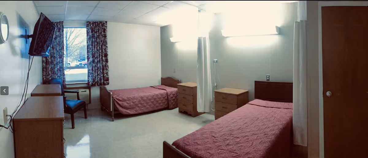 A simple room with two single beds covered in pink bedspreads, two wooden dressers, a chair, a wall-mounted television, and a window with floral curtains. The room has white walls and a tiled floor, with bright overhead lighting.
