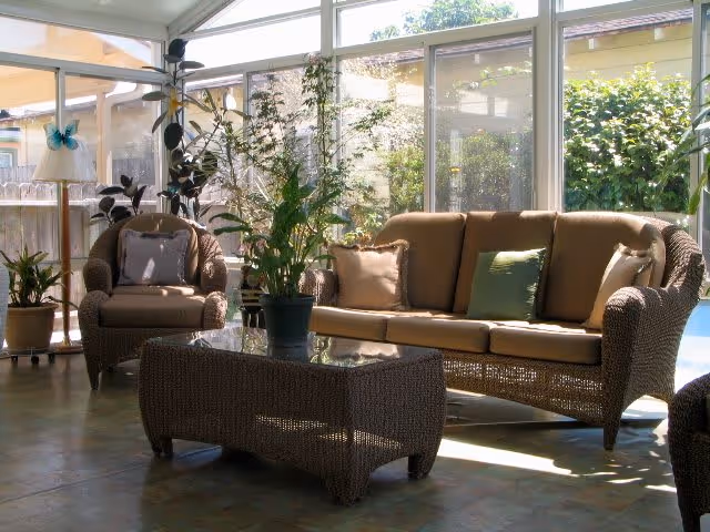 A sunlit indoor seating area with wicker furniture including a sofa and armchair with beige cushions, a glass-top wicker coffee table, several potted plants, and large windows showing an outdoor garden area.