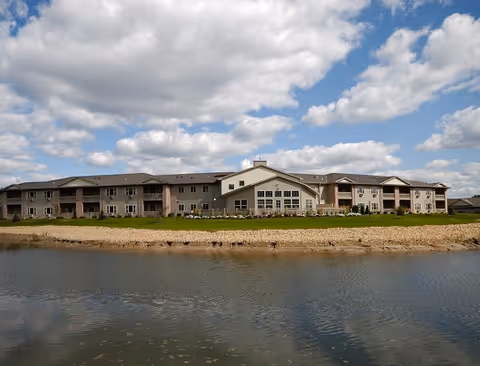 Exterior view of a large two-story senior living facility building with multiple windows and balconies, situated beside a calm body of water under a partly cloudy sky.