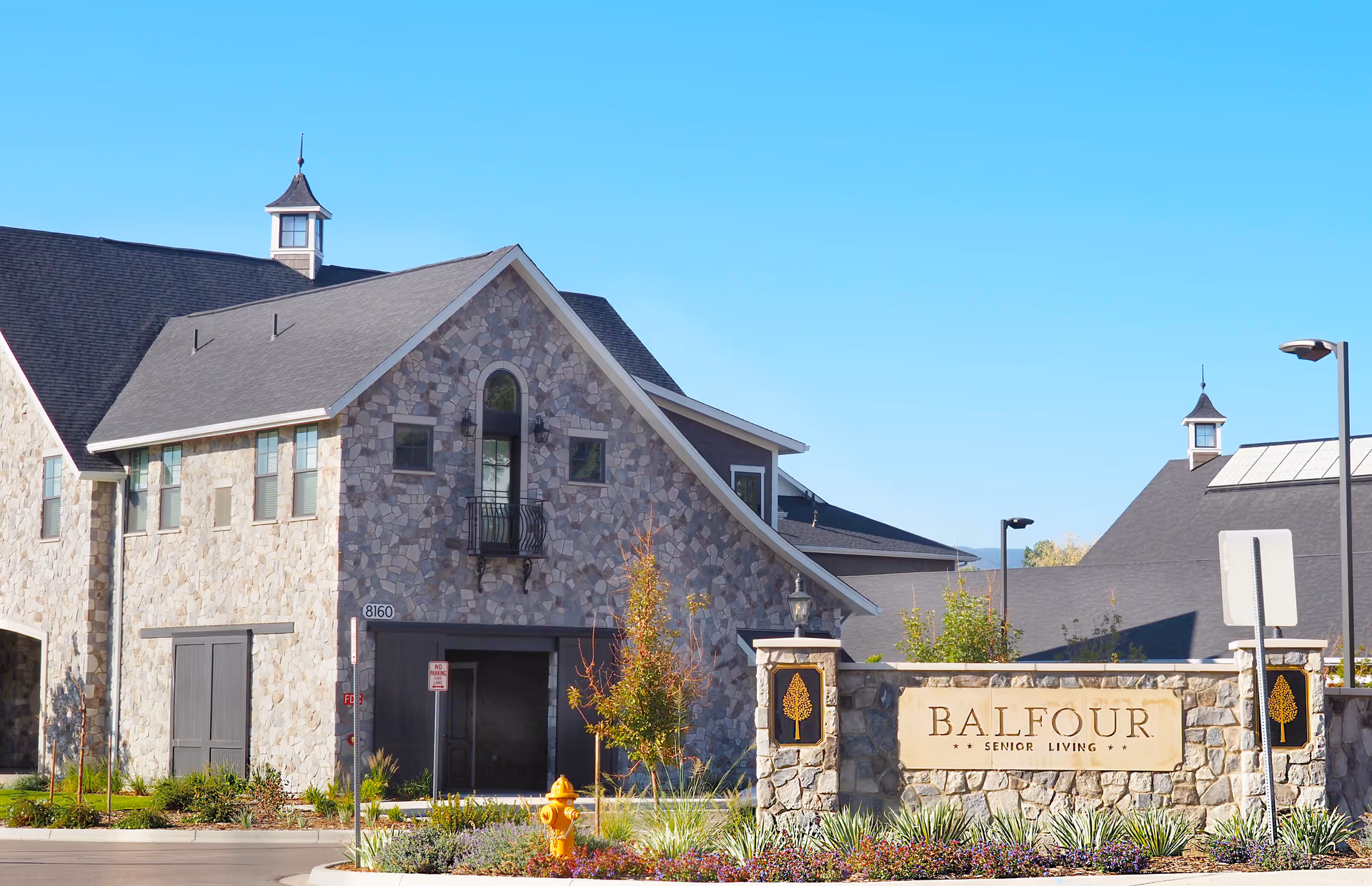 Exterior view of Balfour Senior Living facility featuring stone buildings with peaked roofs, a landscaped area with plants and a fire hydrant, and a stone sign with the name 'Balfour Senior Living' under a clear blue sky.