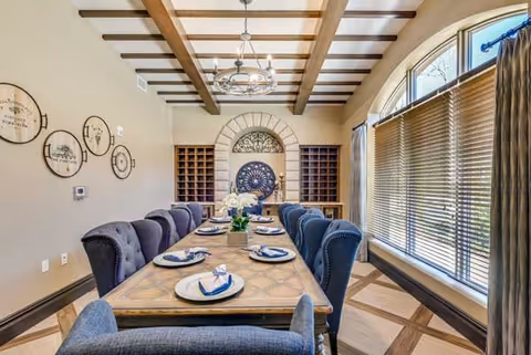A dining room with a long wooden table set with plates and napkins, surrounded by upholstered chairs. The room features wooden ceiling beams, a chandelier, large windows with blinds, and decorative wall art. There are built-in wine racks and a decorative circular wall piece at the far end of the room.