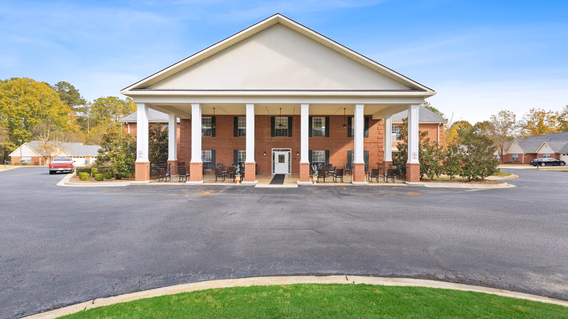 Front exterior view of a two-story brick building with white columns supporting a large covered entrance. There are outdoor seating areas with tables and chairs under the covered entrance. The building is surrounded by a paved driveway and some greenery, with a clear blue sky above.